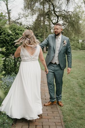 Bride with a bow in her hair and a lace-back white gown dances with her groom in a green suit on a brick path in a spring garden.