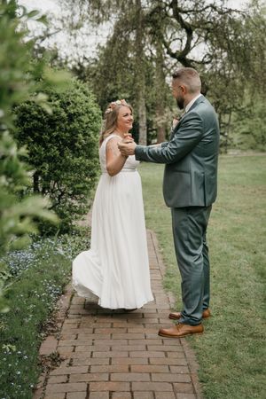 Bride in a flowing white gown and groom in a green suit share a first dance on a brick garden path.