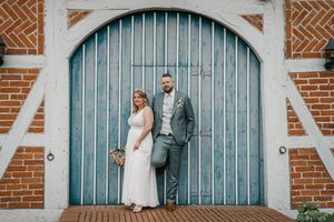 Wedding couple poses in front of a rustic blue barn door with brick and timber framing, holding a bouquet.