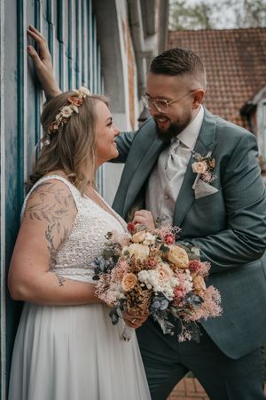 Smiling bride with flower crown and bearded groom in glasses embrace while holding a pastel wedding bouquet.