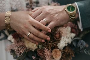Close-up of a couple's hands with wedding rings and gold watches resting over a dried flower bouquet.