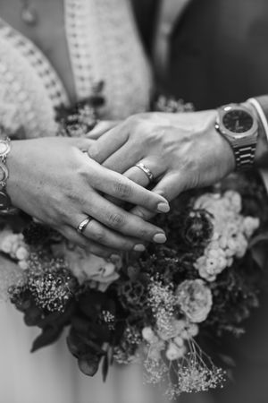 Black and white close-up of newlyweds' hands with wedding rings and watches resting on a floral bouquet.