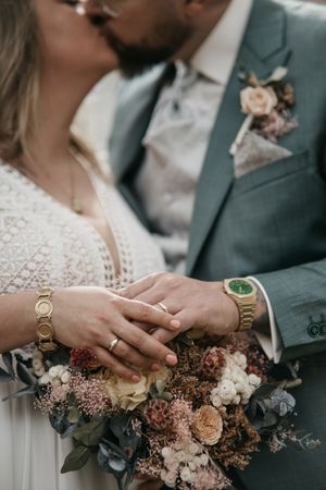 Newlyweds kiss behind a floral bouquet as their ringed hands and gold watches are shown in focus.