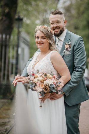 Happy bride with a floral crown and groom in a green suit hug on a tree-lined path, holding a bouquet.