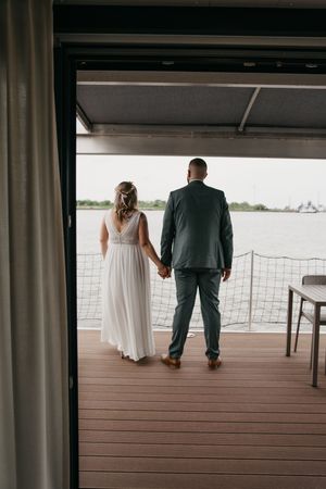 Bride and groom hold hands on a wooden deck, looking out over calm waterfront views on their wedding day.