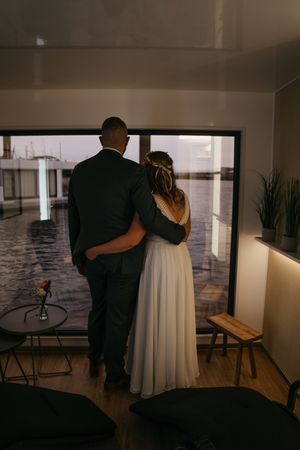 Brautpaar umarmt sich von hinten vor einem großen Fenster mit Blick aufs Wasser in einem gemütlichen Hausboot am Hochzeitstag.