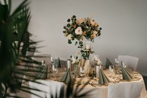 Runder Hochzeitstisch mit floralem Mittelpunkt, salbeigrünen Servietten, Weingläsern und Kerzen, umrahmt von Palmblättern. Lankenauer Höft