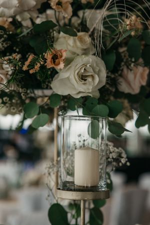 Weiße Rosen, Pfingstrosen und Eukalyptus schmücken einen Glaswindlichtständer mit einer Stumpenkerze auf einer eleganten Hochzeitstafel.