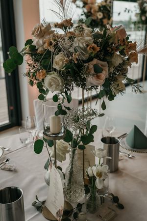 Elegant wedding table centerpiece with cream and peach roses, greenery, a pillar candle and glass vases on a white linen-covered table.