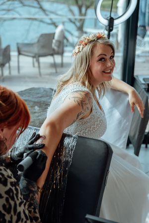 Smiling bride in a white lace gown and floral hair crown gets a tattoo on her arm from a gloved tattoo artist beside a bright window.