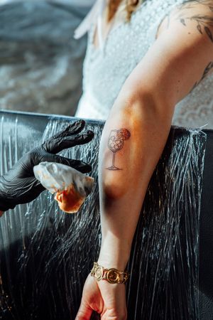 Tattoo artist in black gloves cleans a fresh cocktail glass tattoo on a woman's forearm, with plastic wrap covering the work surface.