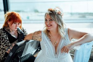 Smiling bride with a flower crown and arm tattoos sits in a white lace dress while a red-haired woman in leopard print works nearby.