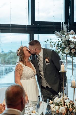 Groom in a gray suit kisses his tattooed bride in a white lace wedding dress beside white floral centerpieces and candles.