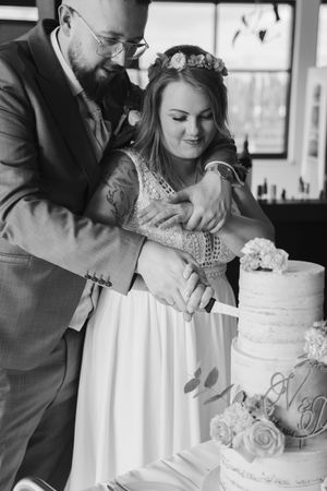 Black and white photo of a bride and groom cutting their rustic tiered wedding cake together, decorated with flowers and initials.