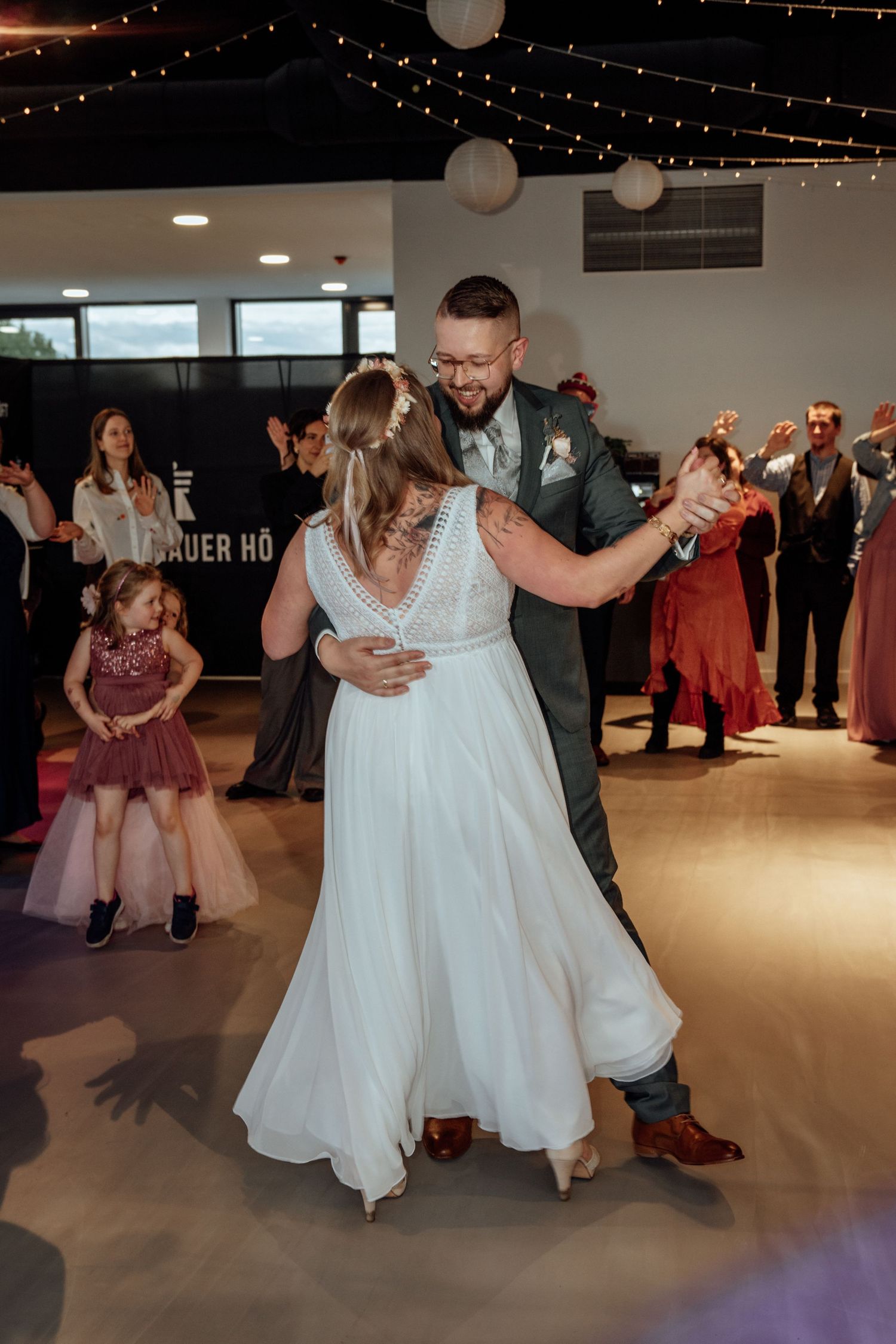 Bride in a flowing white gown and groom in a gray suit share a first dance as guests and a little girl watch from the dance floor.