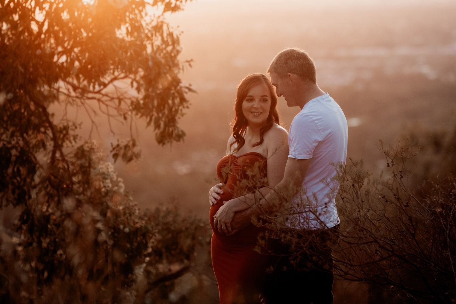 Couple embracing at golden hour sunset on a hilltop, woman in red dress, warm glowing light.