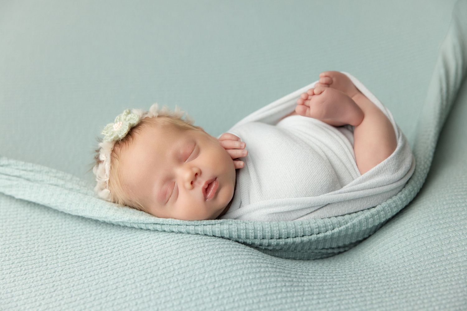 Sleeping newborn baby wrapped in teal swaddle inside a small bucket with a green pillow.