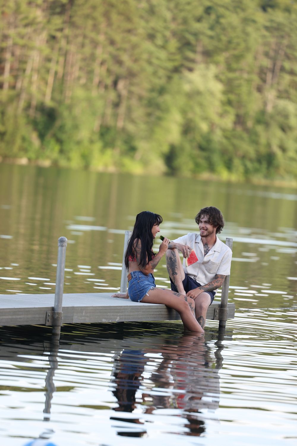 Couple sitting on a wooden dock over a calm lake, sharing drinks and smiling with lush green forest backdrop.