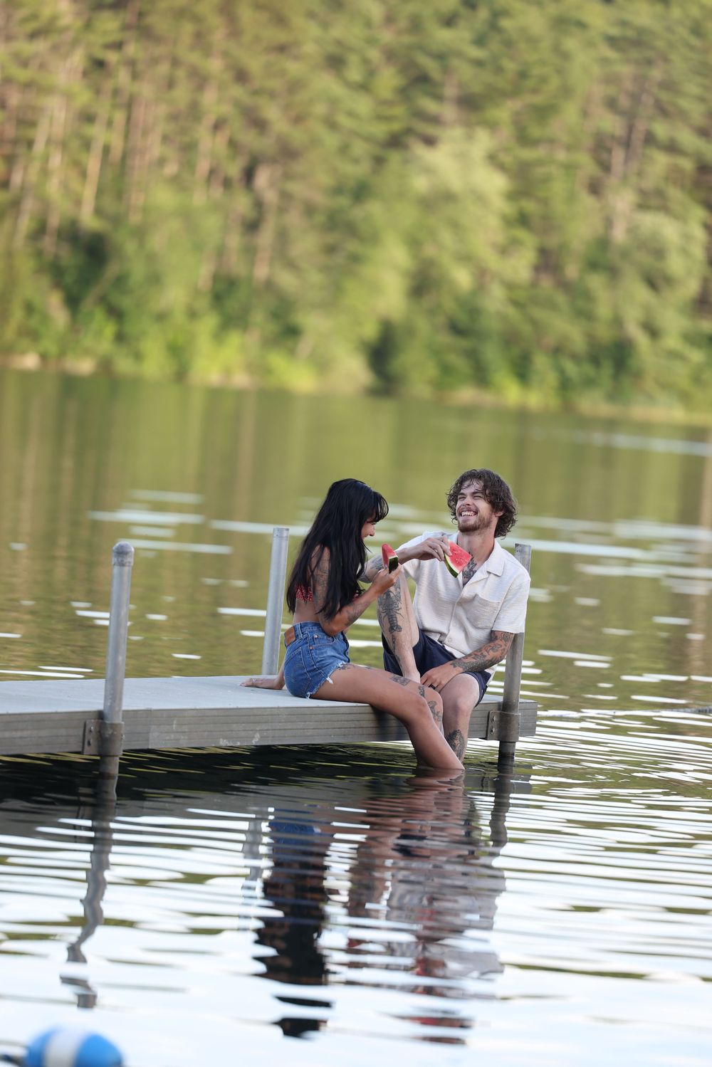 Couple sitting on a lakeside dock sharing watermelon on a sunny summer day.