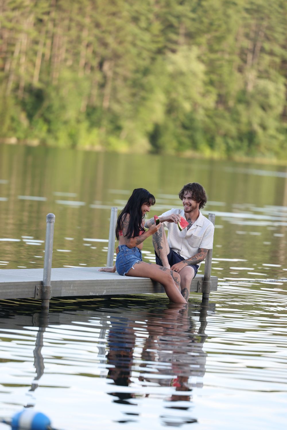 Couple sitting on a wooden dock over a calm lake, sharing watermelon on a sunny summer day.