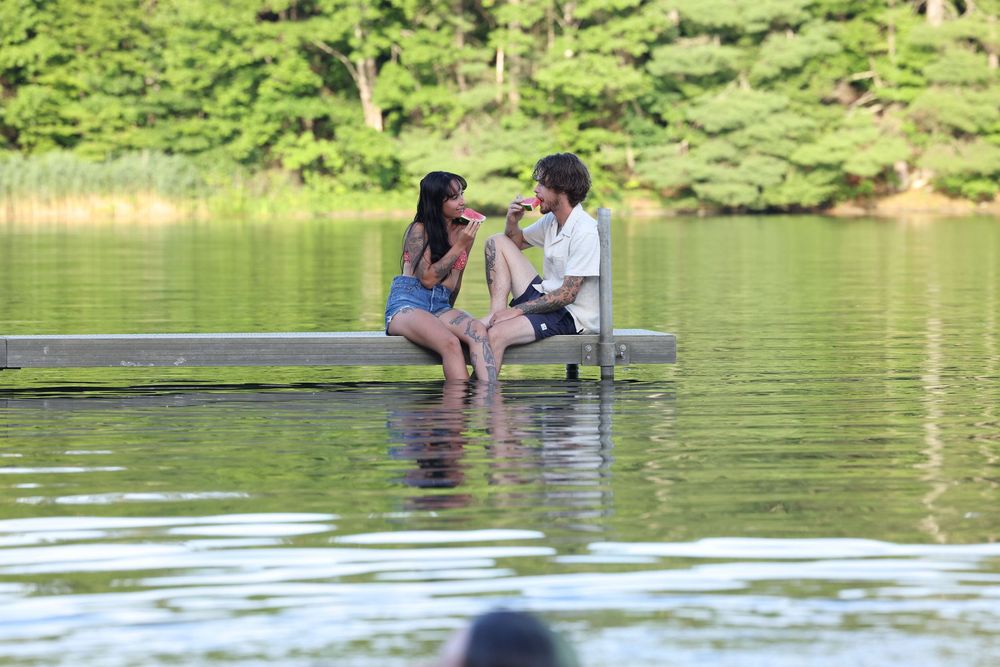 Couple sitting on a dock over a calm green lake, sharing a romantic moment surrounded by lush trees.