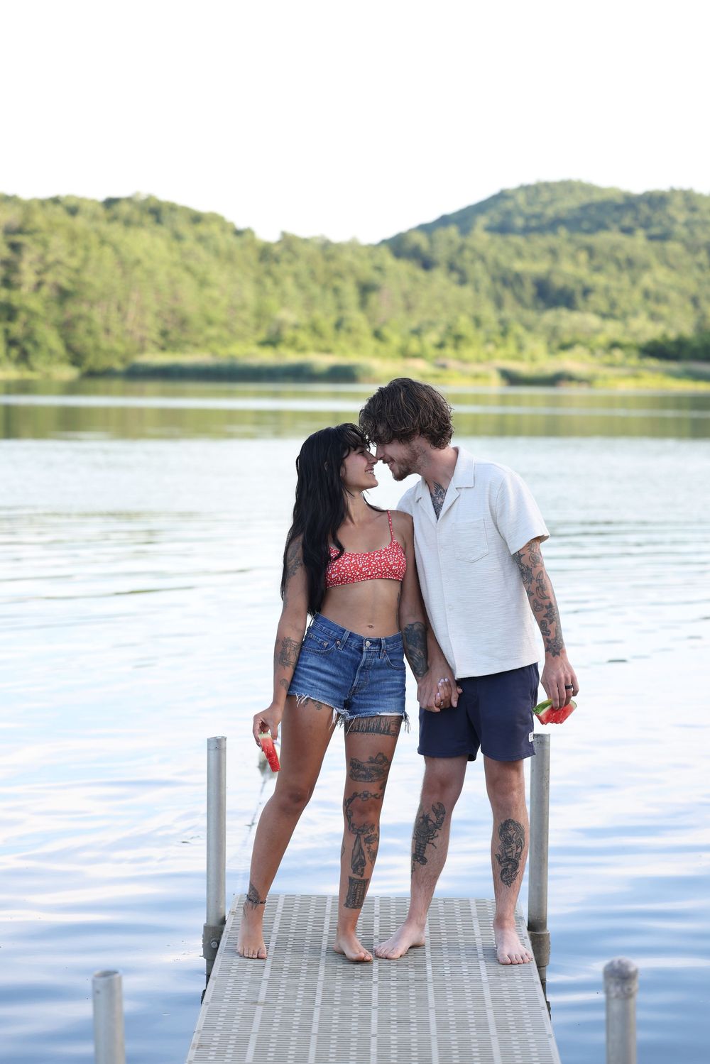 Couple with tattoos kissing on a lake dock, wearing summer clothes, with green hills and calm water in the background.