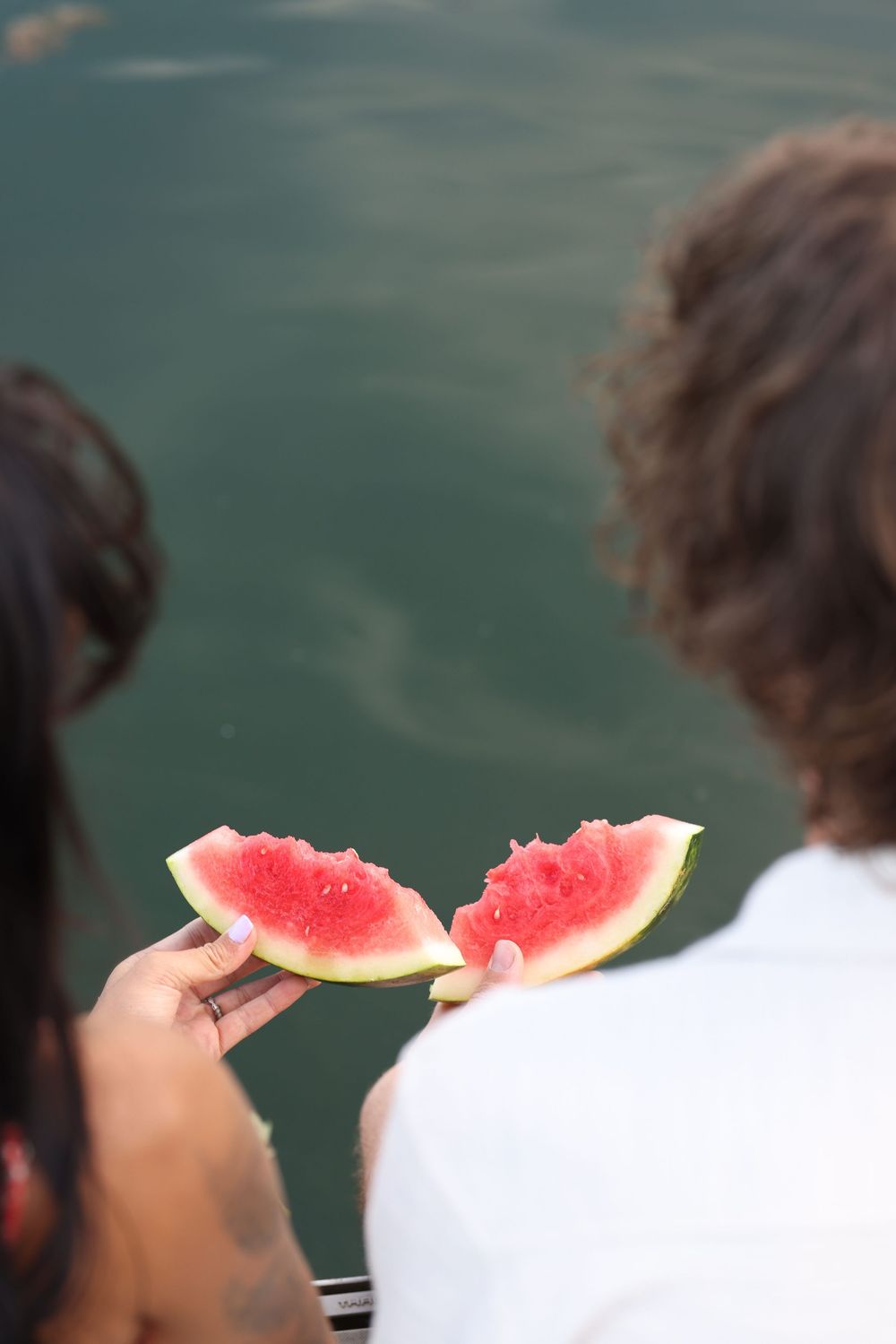 Couple sitting by water sharing two slices of fresh red watermelon on a sunny summer day.