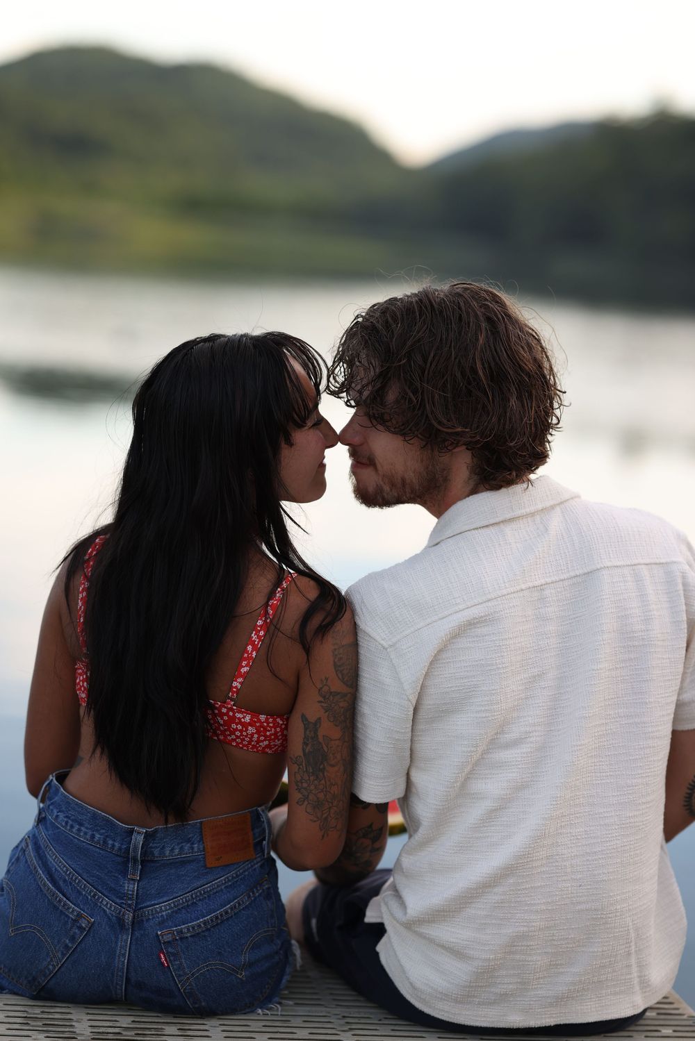 Couple sharing a romantic moment by a lake, sitting on a dock with mountains in the background at dusk.