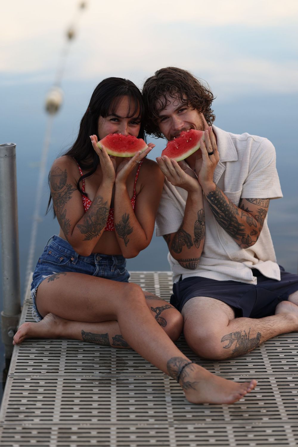 Tattooed couple sitting on a dock eating watermelon slices near a lake at dusk.
