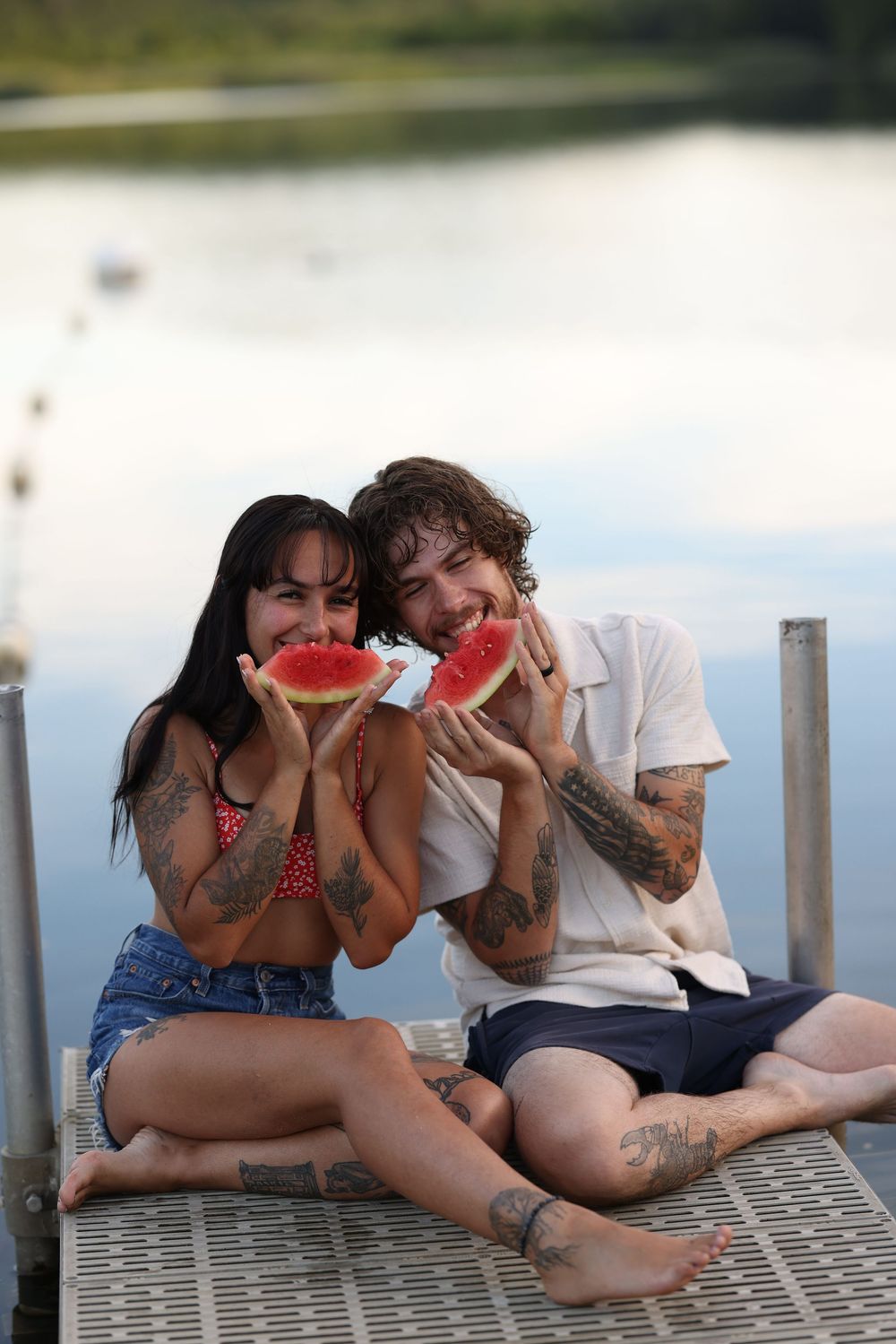 Tattooed couple sitting on a dock eating watermelon slices by a calm lake in summer.