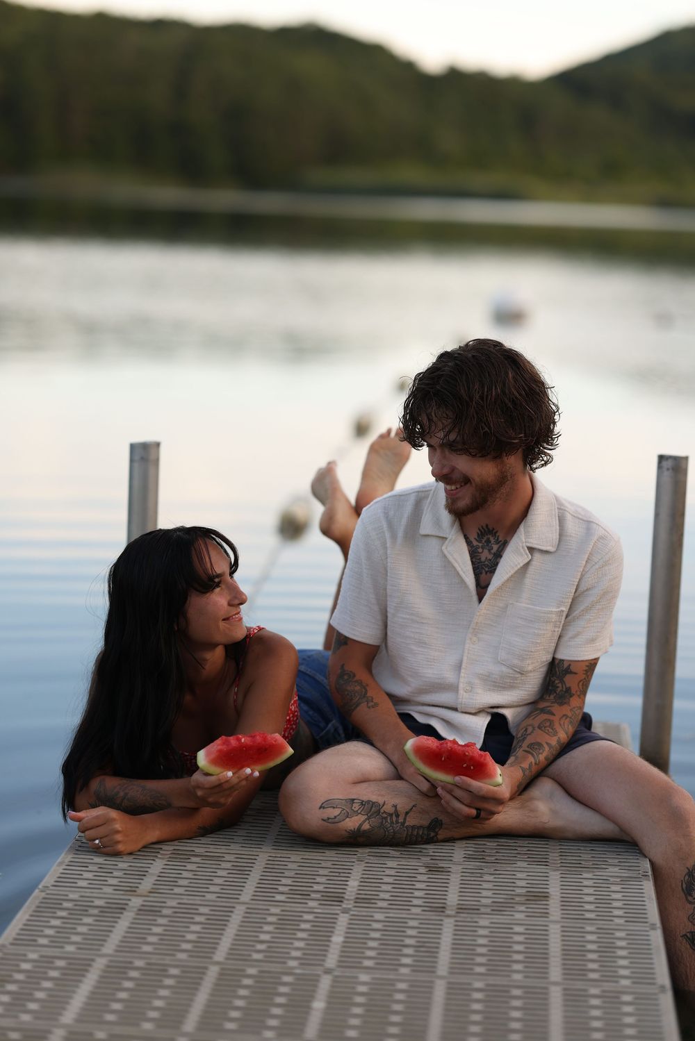 Tattooed couple sitting on a lakeside dock sharing watermelon slices at sunset.