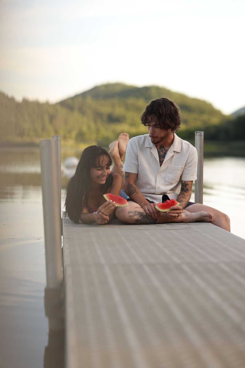 Couple sitting on a dock sharing watermelon slices by a serene lake with green hills in the background.