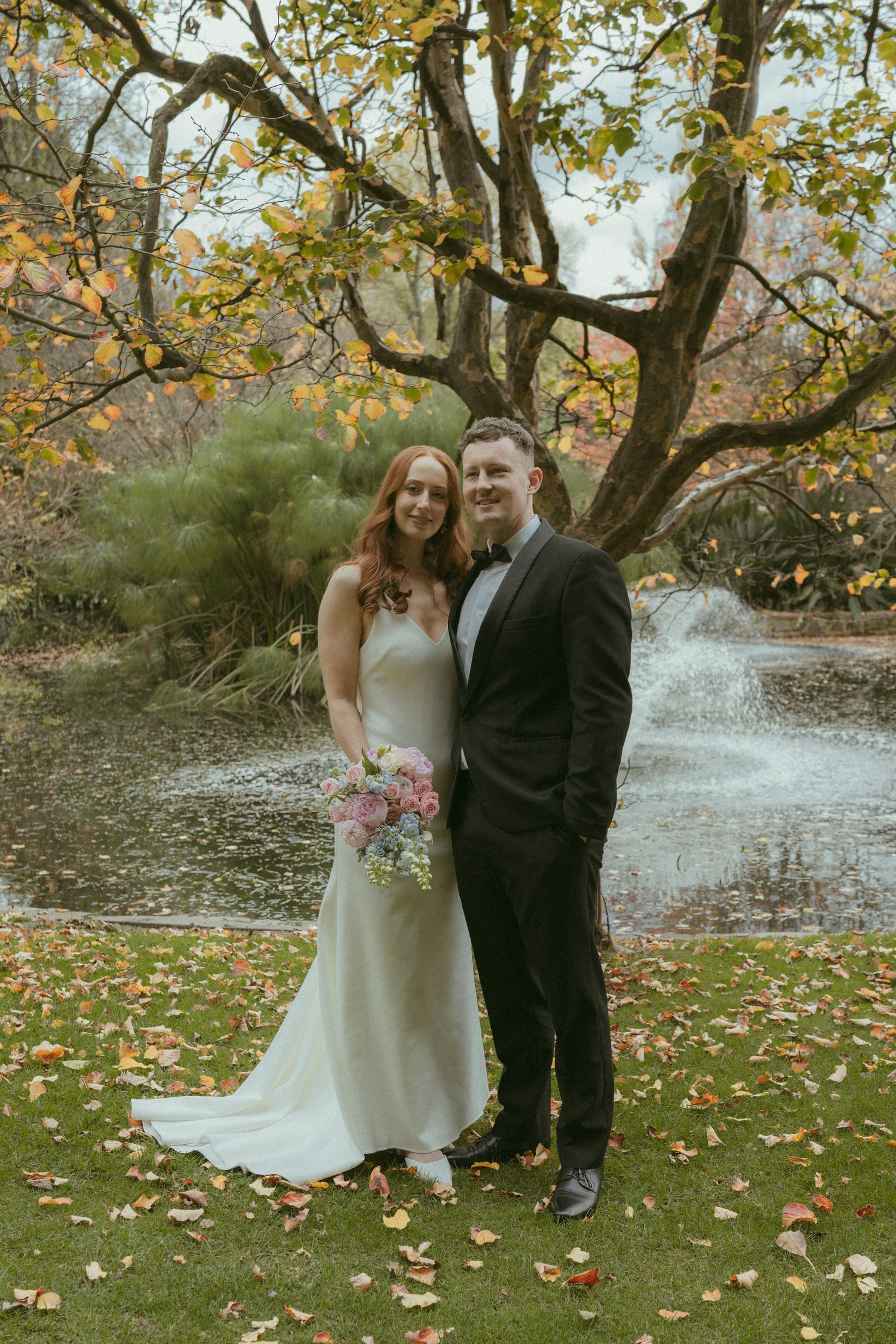 Bride and groom pose by autumn pond with colorful foliage, bride holding pastel bouquet in white gown, groom in black suit.