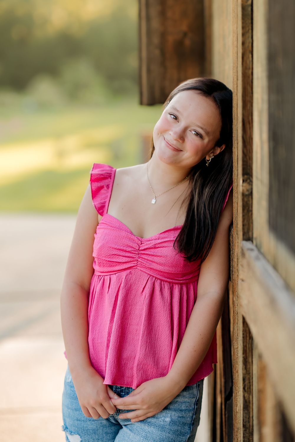 Young woman in pink ruffled top and jeans leaning against rustic wooden wall, smiling outdoors.