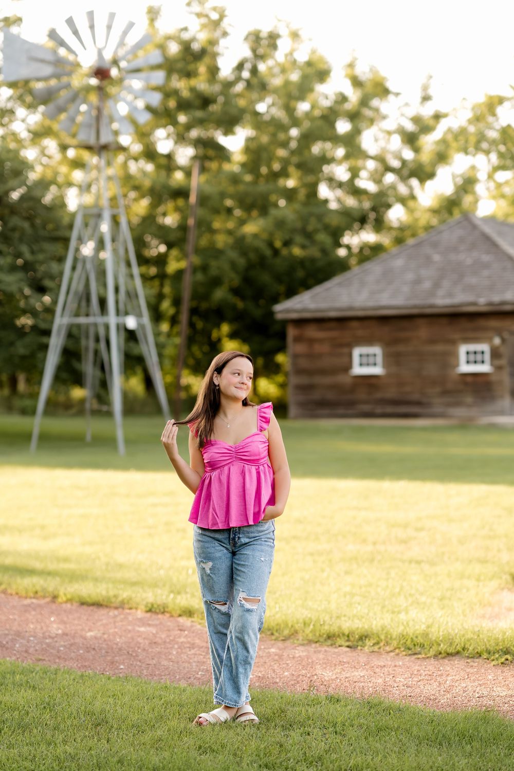 Young woman in pink top and jeans stands near a rustic windmill and wooden barn on a sunny green farm.