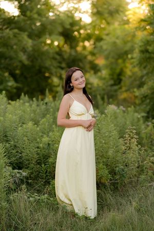 A young girl in a flowing yellow maxi dress smiles while standing in a lush green field at golden hour.