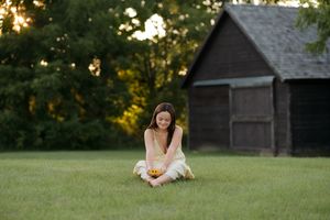Young girl sitting cross-legged on green grass holding yellow flowers near a rustic wooden barn at sunset.