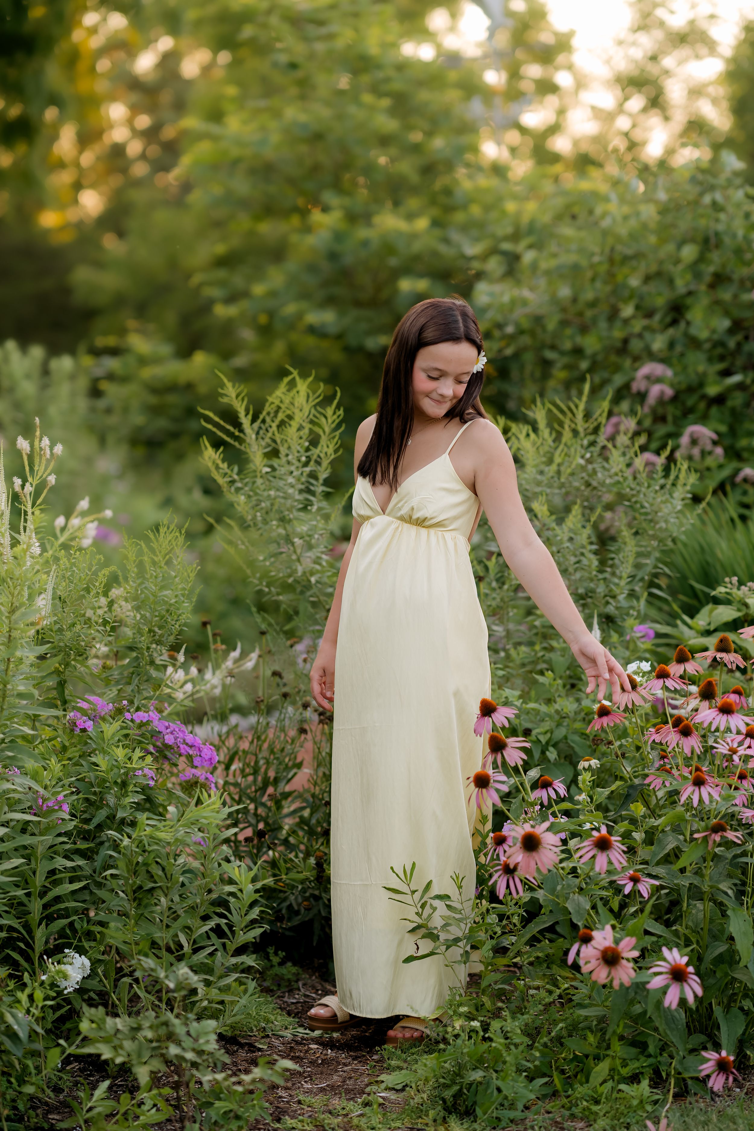 Young woman in yellow maxi dress walking through a lush garden, touching pink echinacea flowers at golden hour.