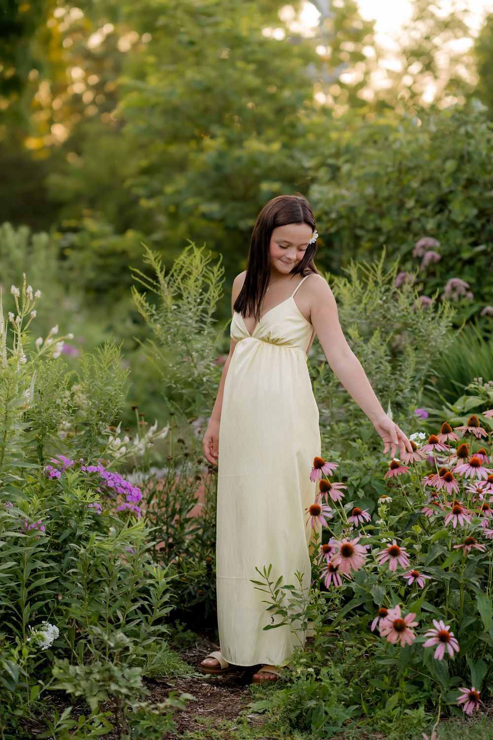 Young woman in yellow maxi dress walking through a lush garden, touching pink echinacea flowers at golden hour.