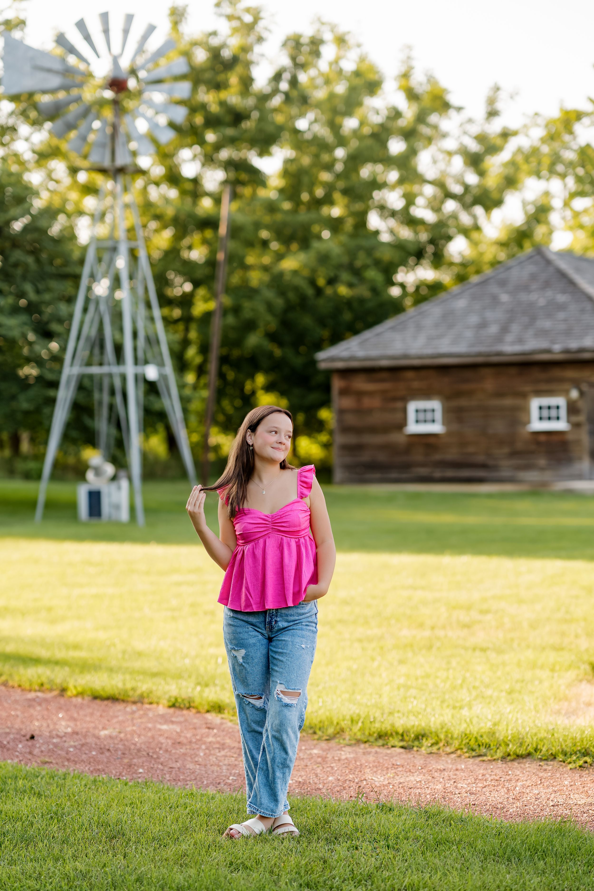 Young girl in pink top and jeans poses near a windmill and rustic barn on a lush green farm.