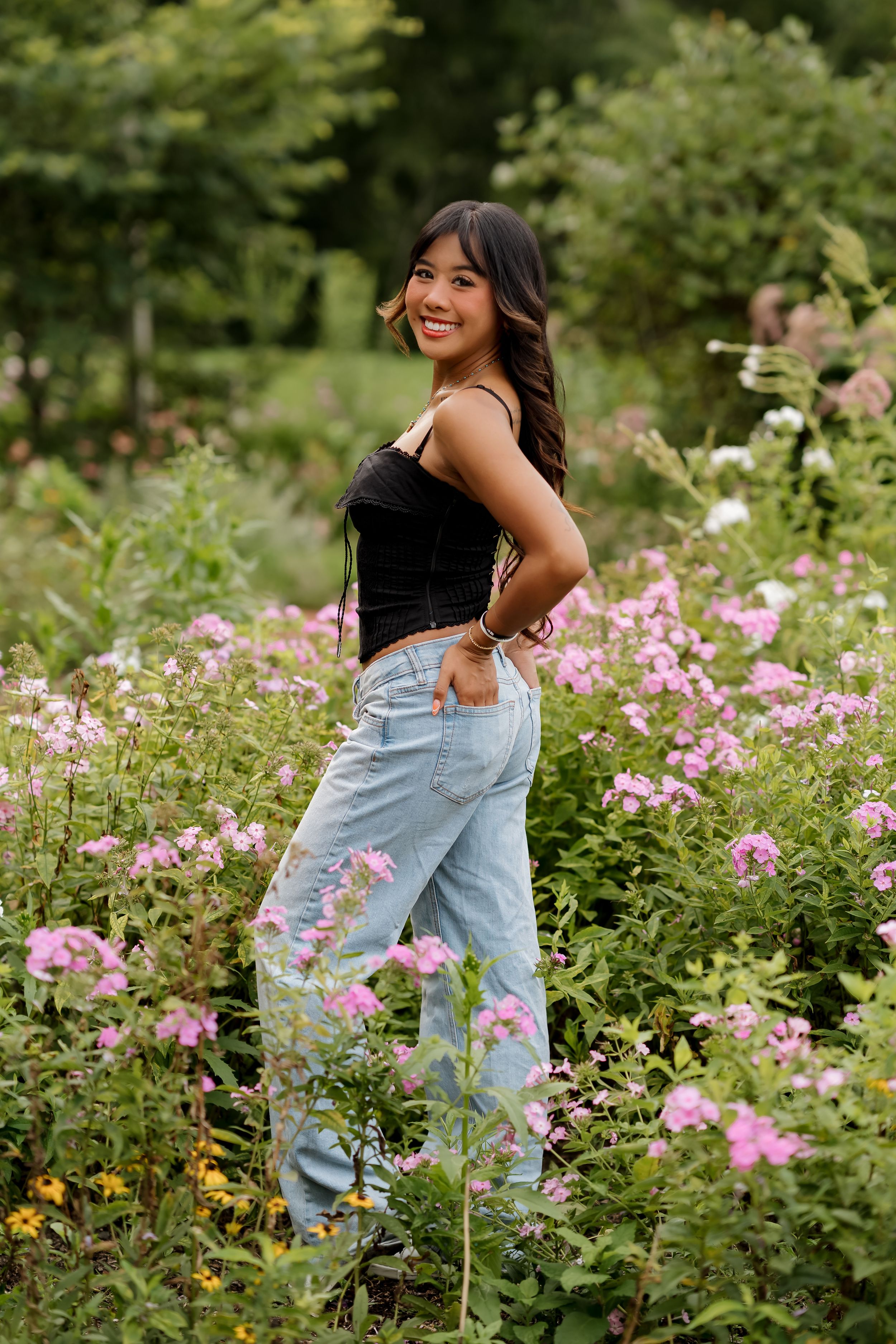 Young woman in black crop top and light blue jeans smiling among pink wildflowers in a lush garden.