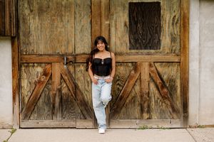 Young woman in black corset top and light jeans leaning against rustic wooden barn doors outdoors.