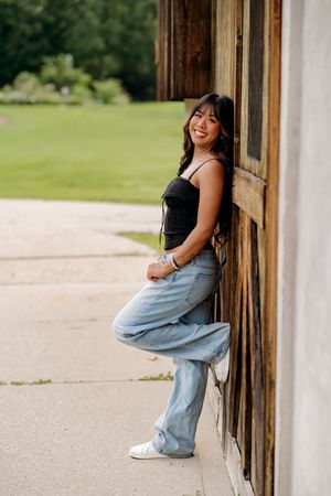 Young woman in black top and wide-leg jeans leans against rustic wooden wall, smiling outdoors.