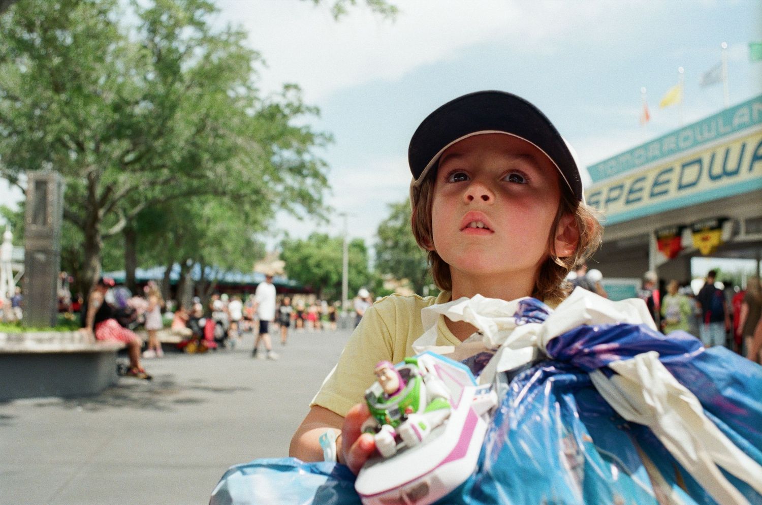 Young boy holding a toy at an outdoor amusement park near a Speedway sign on a sunny day.