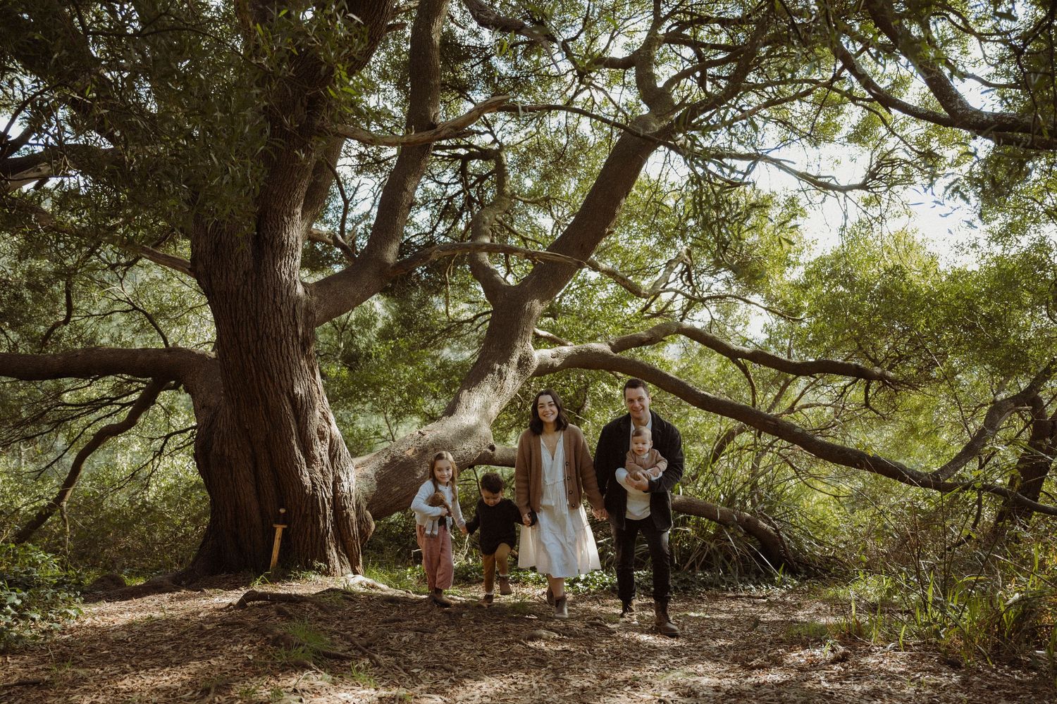 Family of five posing happily in front of a large tree in a lush forest, with children climbing and parents smiling.