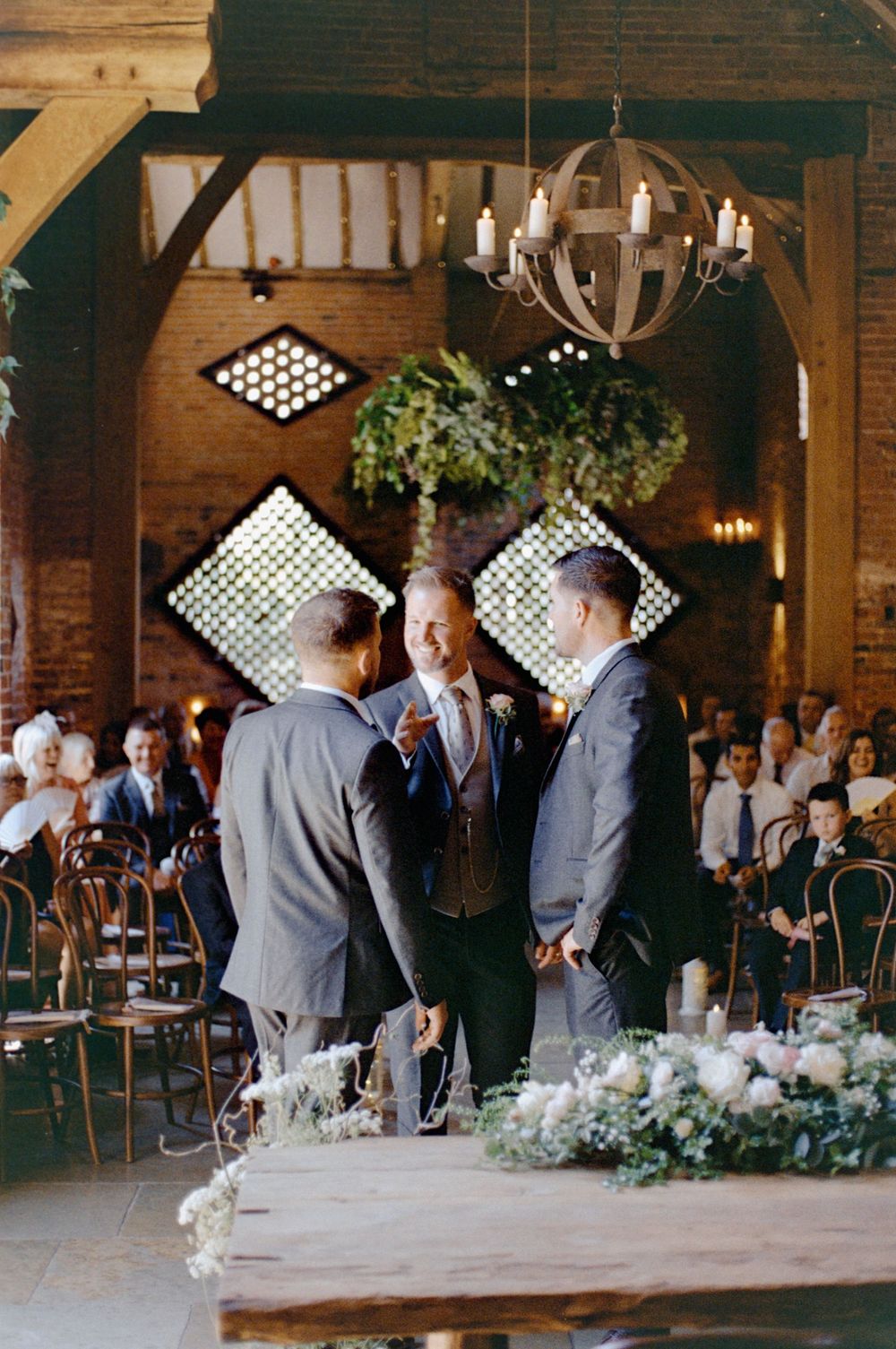 Three groomsmen in grey suits stand at a rustic barn wedding altar adorned with floral arrangements and candlelit chandeliers.