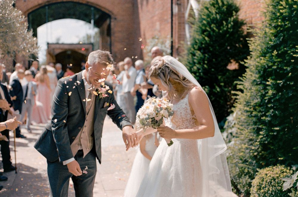 Bride and groom walk through confetti shower at sunny outdoor wedding ceremony near brick archway.