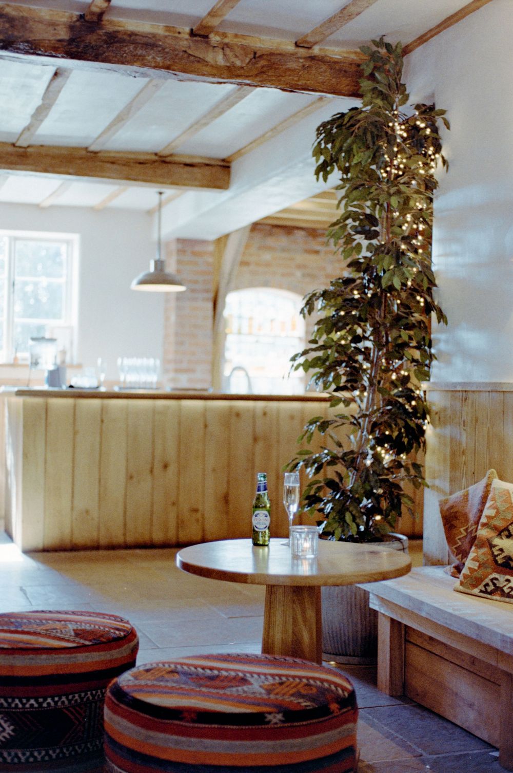 Rustic bar interior with wooden paneling, round table, fairy light tree, striped ottomans, and exposed beam ceiling.