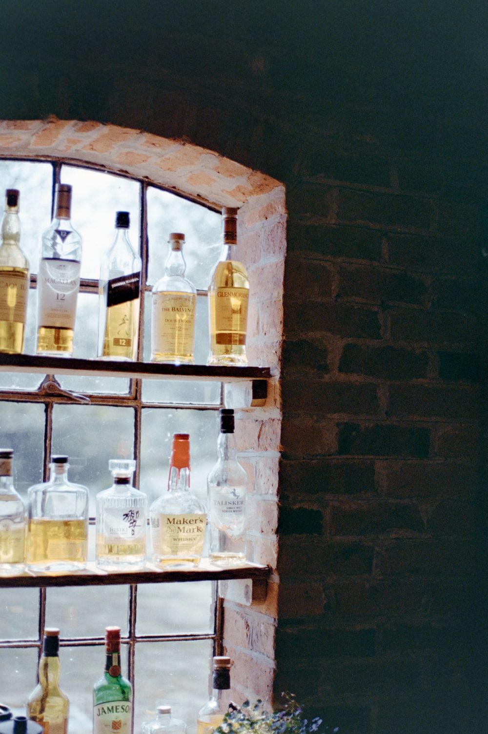 Vintage liquor bottles displayed on rustic brick wall shelves, featuring Maker's Mark and Jameson whiskey in a bar setting.