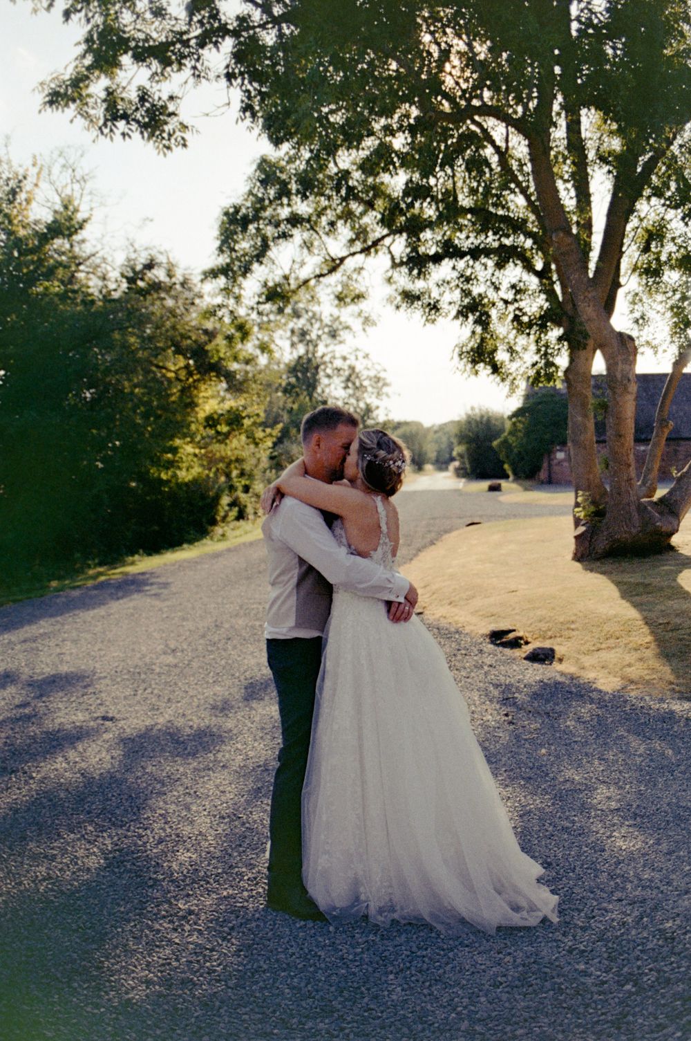 Bride and groom embracing on a gravel path surrounded by lush green trees during golden hour at outdoor wedding.